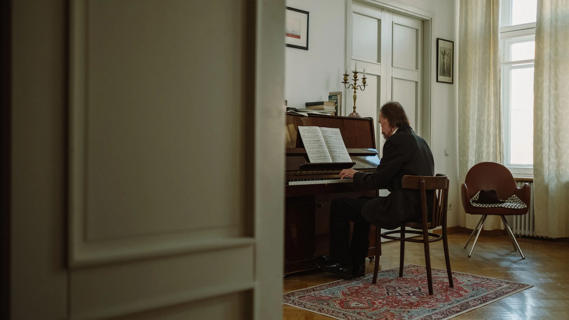 A man in a dark coat plays an upright piano with an open score in a bright, high-ceilinged room with a brass candelabrum, parquet floor and tall windows (not the Chopin Salon).
