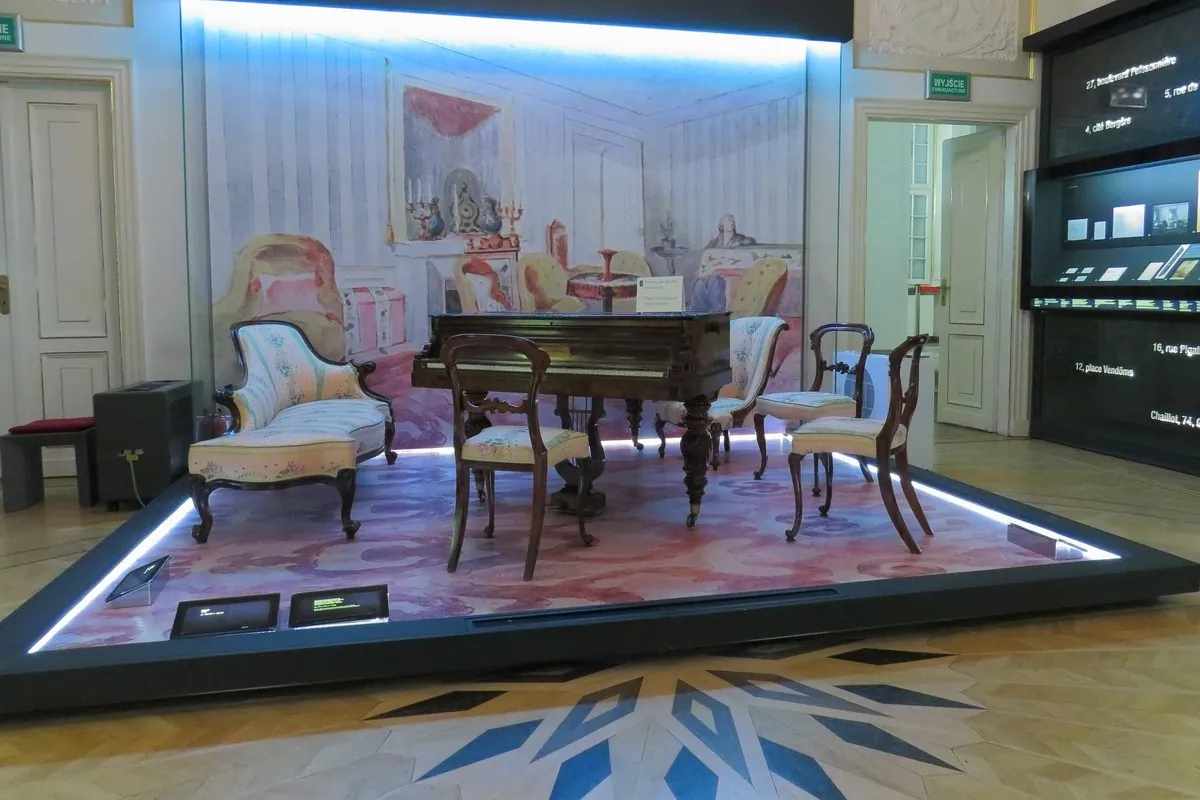 A small grand piano with a score on the desk stands among chairs and a sofa in a lit museum display set like a drawing room